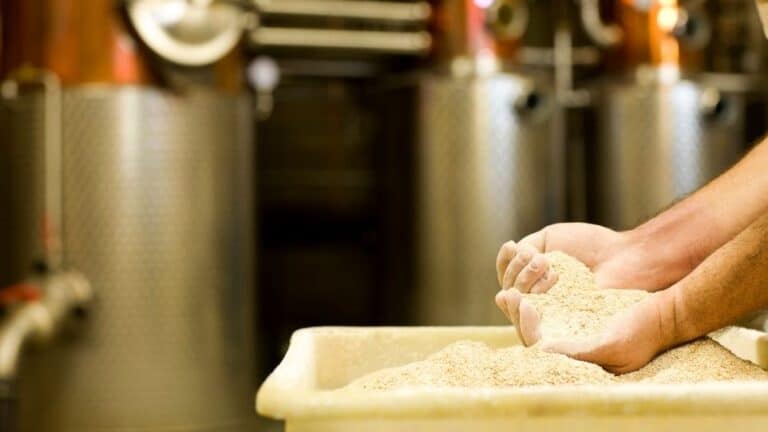 hands of a man in the mash at a distillery with the distilling equipment in the background